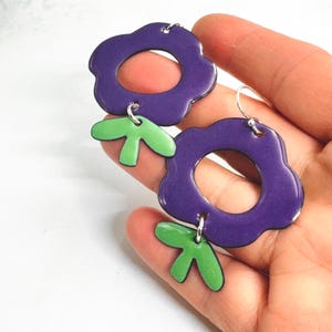 May include: Earrings featuring a purple flower design with green leaf details. The earrings have a glossy finish and are suspended from silver-coloured hooks. The floral design is a bold, retro style, suitable for adding a pop of colour to any outfit.