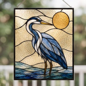 May include: Stained glass panel featuring a blue heron standing in water, with a golden sun in the background. The artwork is framed in black and hangs from a chain. The heron has blue and white feathers and a yellow beak.