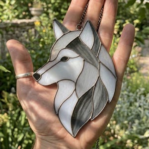 May include: A stained glass suncatcher shaped like a wolf head in profile, featuring white and grey marbled glass segments joined by dark metal solder lines. The piece includes a small metal loop at the top for hanging with a thin metal chain.
