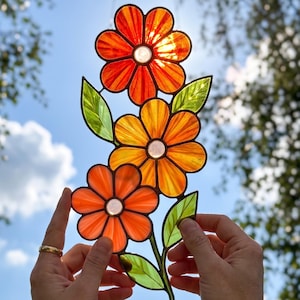 May include: A stained glass floral art piece featuring three orange and yellow flowers with green leaves. The sun shines through the glass, creating a vibrant display. The piece is held up against a blue sky.