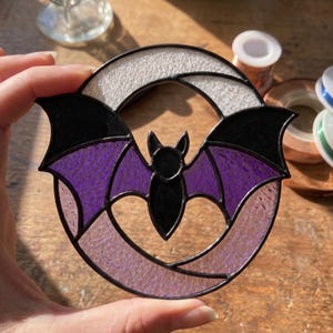 May include: A circular stained glass suncatcher featuring a black bat with purple wings centred against a white crescent moon. The piece is constructed with dark metal solder lines separating the textured glass segments in black, purple, and white.