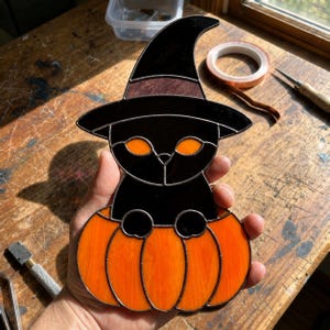 May include: A stained glass suncatcher featuring a black cat with glowing orange eyes wearing a pointed black witch hat, sitting inside a bright orange pumpkin. The piece is constructed with dark metal solder lines connecting the glass segments.