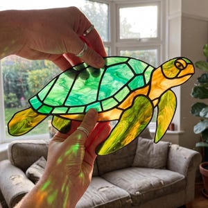 Op de afbeelding: Een gebrandschilderde schildpad met een groen en geel kleurenschema. De schild van de schildpad is een mozaïek van groen glas, terwijl de kop, vinnen en randen geel zijn. Het glas wordt door handen vastgehouden.