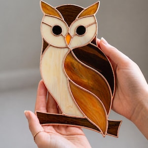 May include: A stained glass owl suncatcher featuring cream, amber, and dark brown glass pieces joined by copper foil solder lines. The owl is depicted perched on a horizontal branch, with two black glass eyes and a small orange glass beak.
