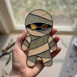 May include: A stained glass mummy figure constructed from cream and sage green textured glass pieces joined by dark solder lines. The character features two almond-shaped eyes in amber and orange glass set against a black glass background.