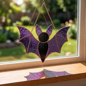 May include: A stained glass bat suncatcher featuring textured purple wings and a solid black body. The bat is framed in copper-colored metal and hangs from a matching copper-colored metal chain, casting a purple shadow on the wooden surface below.