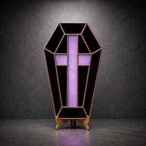 May include: A coffin-shaped stained glass panel featuring opaque black glass segments surrounding a central lavender purple cross. The glass pieces are joined by copper-colored metal solder lines, and the entire piece is supported by a gold-toned stand.