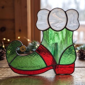 May include: A stained glass elf shoe ornament featuring a white cuff, a green body, and a red toe and heel. The shoe has a curled toe with a small metal bell attached. The glass pieces are joined by dark metal solder lines throughout the design.