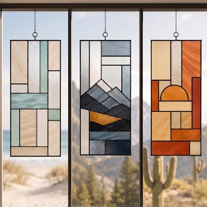 Puede incluir: Tres paneles rectangulares de vidrieras colgados de cadenas. El panel izquierdo presenta formas geométricas en beige y azul claro. El panel central representa un paisaje montañoso en azul, gris y dorado. El panel derecho muestra una puesta de sol en naranja y beige.