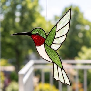 May include: A stained glass hummingbird suncatcher featuring a vibrant red throat, green wings, and a white underbelly. The bird is constructed with individual glass segments held together by dark metal solder lines, suspended by a thin clear filament.