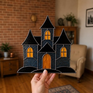 May include: A stained glass haunted house decoration featuring dark blue and black textured glass panels with bright orange glass windows and a central arched orange door, all held together by thin dark metal lead lines in a castle shape.