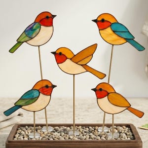 May include: Five stained glass bird ornaments on thin metal rods. Each bird features a cream-colored chest, a red face, and wings in varying shades of green, blue, and orange. The birds are mounted on clear circular bases and arranged in a group.