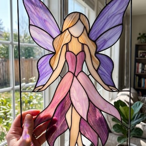 Pode incluir: Obra de arte em vitral retratando uma fada com asas e vestido roxo e rosa. A fada tem cabelo loiro e está sobre um fundo transparente. A obra de arte é emoldurada com metal preto.