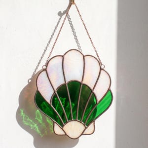 May include: A stained glass suncatcher shaped like a seashell, featuring iridescent white glass panels at the top and deep green glass panels at the bottom, all held together by dark metal solder lines and suspended by a thin metal chain.