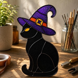 Op de afbeelding: Een glas-in-loodfiguur van een zwarte kat met een paarse heksenhoed met een oranje band en een gele gesp. De kat heeft gele ogen en is opgebouwd uit gestructureerde zwarte glasstukken die verbonden zijn door donkere metalen soldeerlijnen.