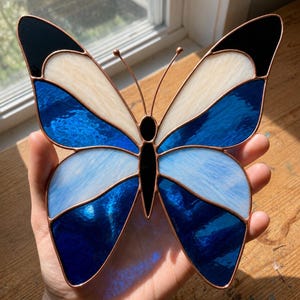 May include: A stained glass butterfly suncatcher featuring a symmetrical design with deep blue, light blue, cream, and black glass segments held together by thin copper foil lines, including two thin wire antennae extending from the central body.