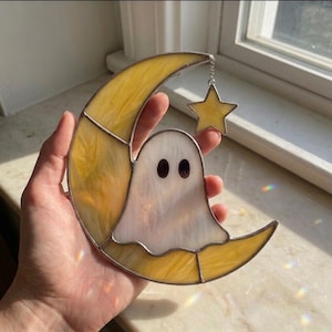 Pode incluir: Um apanhador de sol em vitral com uma lua crescente amarela e um fantasma branco com dois olhos ovais escuros. Uma pequena estrela de vitral amarelo pende do topo da lua por uma corrente prateada. A peça é construída com linhas de solda metálicas.