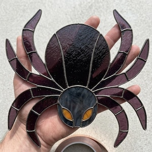 May include: A stained glass spider ornament featuring a large, dark purple abdomen and a smaller, textured grey head. The spider has eight curved legs made of deep purple glass segments joined by dark metal solder lines and two bright orange eyes.