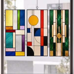 Pode incluir: Três painéis retangulares de vitral pendurados por correntes. O painel esquerdo apresenta uma grade de vidro amarelo, azul, verde, vermelho e roxo. O painel central exibe um círculo amarelo e formas geométricas em vermelho, azul e preto. O painel direito mostra vidro verde e dourado em um padrão vertical.