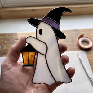 Op de afbeelding: Een glas-in-lood figuur van een geest met een wit lichaam, een zwarte heksenhoed met een paarse band en een kleine amberkleurige lantaarn. De geest heeft één zwart oog en is afgewerkt met donkere metalen soldeerlijnen langs de randen.