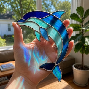 Op de afbeelding: Een glas-in-lood dolfijn suncatcher met een springend ontwerp, bestaande uit segmenten van diep marineblauw, koningsblauw en doorschijnend licht turkoois glas, verbonden door dunne zilverkleurige metalen soldeerlijnen voor een levendig effect.