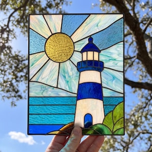 May include: Stained glass panel featuring a lighthouse with blue and white stripes, a blue dome, and a golden sun. The artwork depicts a coastal scene with blue water and green foliage. The panel is held against a backdrop of a blue sky and tree branches.