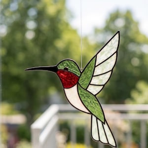 Op de afbeelding: Een glas-in-lood kolibrie zonnevanger met een levendige rode keel, groene vleugels en lichaam, en witte vleugel- en staartveren. De vogel is geconstrueerd met donkere metalen soldeerlijnen en hangt aan een dunne, doorzichtige monofilament draad.