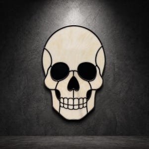 Puede incluir: Un adorno de pared de vitral con forma de cráneo, compuesto por segmentos de vidrio veteado en color blanco roto unidos por líneas de plomo negro. El diseño incluye inserciones de vidrio negro sólido en las cuencas oculares y la nariz.