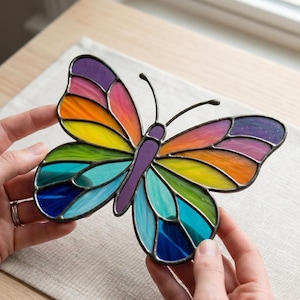 Puede incluir: Una mariposa de vitral con un diseño de arcoíris de colores vibrantes. Las alas están compuestas por segmentos individuales de vidrio en tonos morado, rosa, naranja, amarillo, verde, turquesa y azul, unidos por líneas de soldadura oscuras.