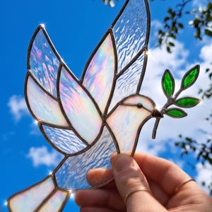 May include: Stained glass dove with iridescent wings and a green olive branch in its beak. The dove is made of clear and white glass, with a brown border. The background is a bright blue sky with some tree branches.