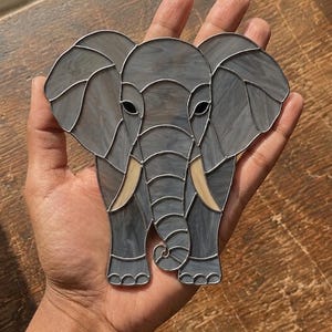 May include: A stained glass elephant ornament featuring grey marbled glass panels joined by thin metal solder lines. The elephant has large ears, two curved cream-coloured tusks, and a curled trunk, with small black glass pieces for the eyes.