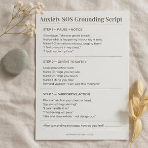 May include: A white card with the text "Anxiety SOS Grounding Script" and instructions for managing anxiety. The card is surrounded by dried flowers and a smooth gray stone. The script includes steps for pausing, noticing, orienting to safety, and supportive action.