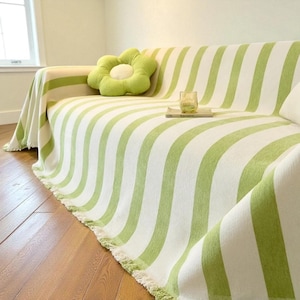 May include: A sofa draped with a green and white striped throw blanket. A green flower-shaped pillow and a glass of water sit on the sofa. The throw has a fringed edge. The sofa is on a hardwood floor.