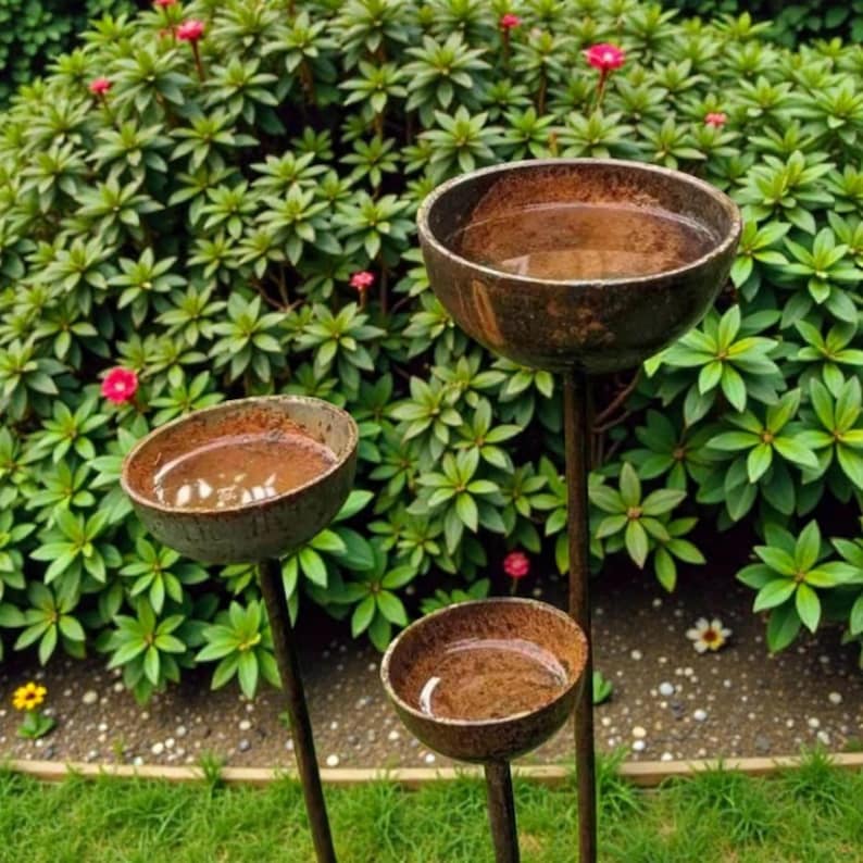 May include: Three rustic, metal bird baths of varying heights stand in a garden. Each bath has a shallow, bowl-shaped basin filled with water. The metal has a weathered, brown finish, contrasting with the green foliage and pink flowers in the background.