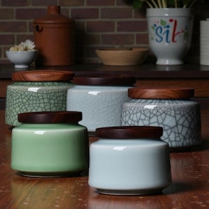 May include: A selection of ceramic jars with wooden lids. The jars are in shades of green and blue, featuring a crackle glaze. The jars are displayed on a wooden surface.