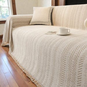 May include: A beige sofa with a matching throw blanket and pillow. The blanket has a textured chevron pattern. A white teacup and saucer sit on the sofa, along with a small book.