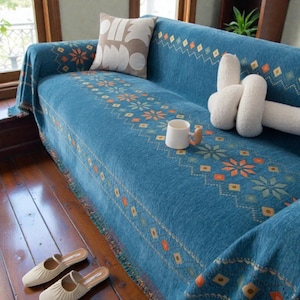 Può includere: Un copridivano blu con un motivo geometrico e floreale nei toni dell'arancione, giallo e verde acqua. La coperta presenta un bordo sfrangiato ed è decorata con un cuscino beige con forme bianche e un cuscino decorativo bianco a nodo.