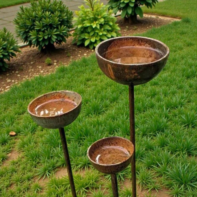 May include: Three rustic, metal bird baths of varying sizes stand in a grassy yard. Each bowl-shaped bath is filled with water and mounted on a slender, dark brown pole. The bird baths are surrounded by green grass and small bushes.