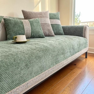 Puede incluir: Un sofá verde con cojines a juego. El sofá tiene una tela texturizada y tejida, con un ribete de color claro. Los cojines son en tonos verdes y grises, con un diseño texturizado. Una taza y un platillo están sobre el sofá.