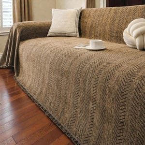 May include: A brown sofa with a herringbone pattern throw. A white cushion with a dark border and a decorative knot cushion are on the sofa. A white teacup and saucer sit on a small book.