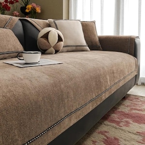 Könnte beinhalten: Ein Sofa mit einem strukturierten hellbraunen Stoffbezug und dunkelbraunen Zierleisten an den Kanten. Das Sofa umfasst mehrere quadratische beige Kissen und ein rundes, mehrfarbiges geometrisches Kissen. Eine weiße Tasse steht auf Papier.