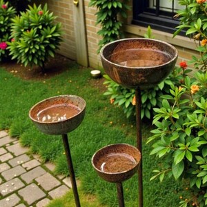 May include: Three rustic, metal bird baths of varying heights stand in a garden. Each bird bath has a shallow, bowl-shaped basin filled with water. The metal has a weathered, brown finish, and the bird baths are set against a backdrop of green foliage and a brick pathway.