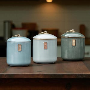 May include: Three ceramic jars in blue, white, and green. Each jar has a rounded lid and a small, rectangular label. The jars have a smooth finish and are arranged on a dark wooden surface. The lids are topped with a gold-coloured handle.