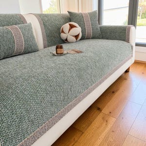 Peut inclure: Un canapé blanc avec un tissu vert texturé et des coussins assortis. Le canapé a une housse verte avec une bordure marron et blanche. Une boule décorative et une petite bougie sont posées sur le canapé.