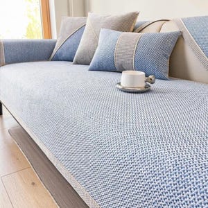 Puede incluir: Una funda de sofá con estampado azul y blanco con cojines a juego. Los cojines tienen detalles en azul y gris. Una taza de té blanca está sobre un platillo plateado en el sofá.