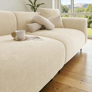 May include: A cream-colored sofa with a textured herringbone fabric pattern. The sofa features a rounded armrest, a matching back cushion, and a soft, curved throw pillow. A white ceramic mug sits on a small saucer on the seat cushion.