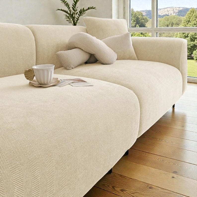 May include: A cream-colored sofa with a textured herringbone fabric pattern. The sofa features a rounded armrest, a matching back cushion, and a soft, curved throw pillow. A white ceramic mug sits on a small saucer on the seat cushion.