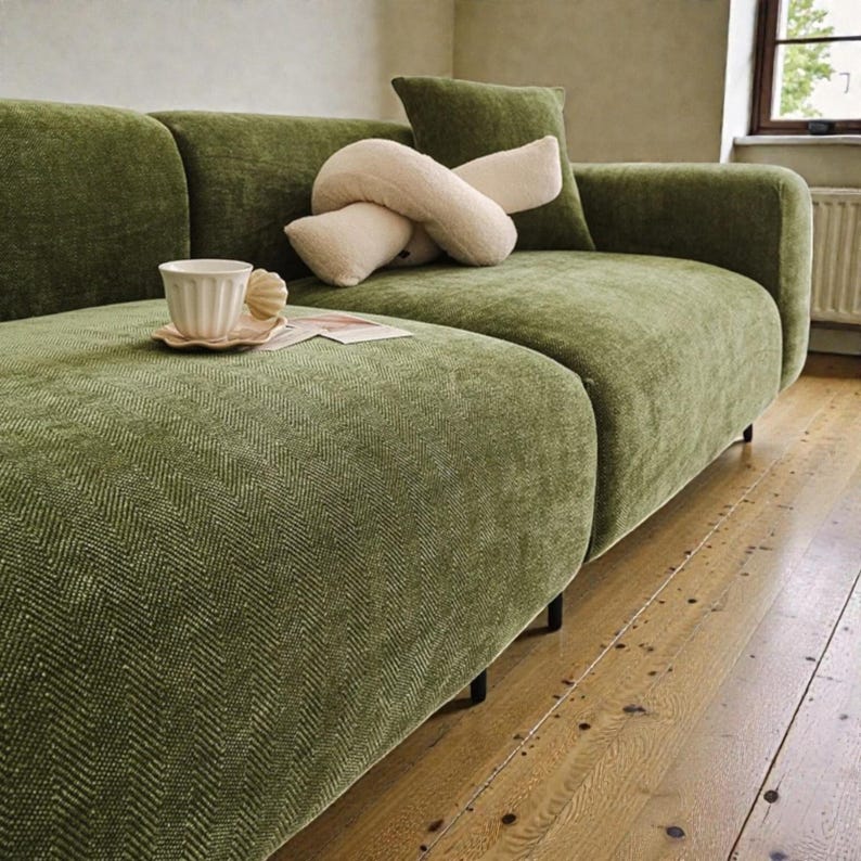 May include: A moss green sofa with a textured herringbone fabric pattern. A cream-colored knot pillow rests on the seat cushion. A small, fluted ceramic cup sits on a matching saucer on the sofa, accompanied by a small rectangular paper card.