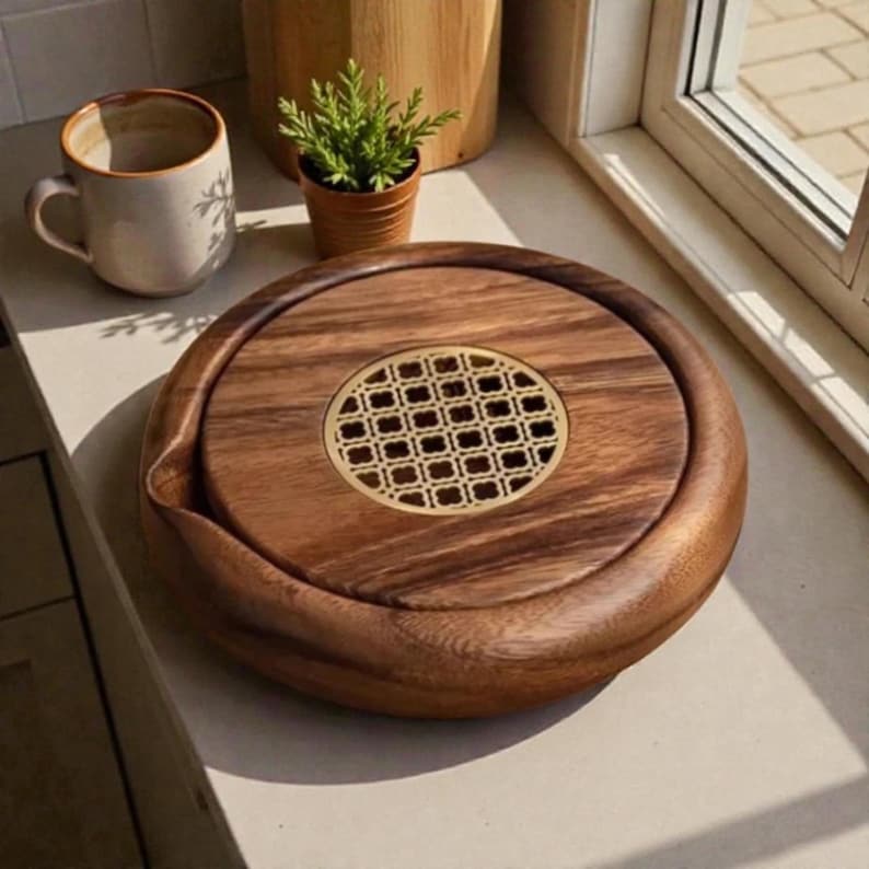May include: A round wooden tea tray with a decorative brass-colored center. The tray has a dark wood grain and a small spout. A ceramic mug and a small potted plant are in the background.