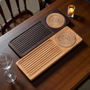 以下が含まれることがあります： 木製のティートレイ2つ。濃い茶色と薄い茶色で、織りのコースターが付いています。濃い茶色のトレイはリブのデザインで、薄い茶色のトレイはスラットのデザインです。ガラスホルダーに入ったろうそくが近くにあります。